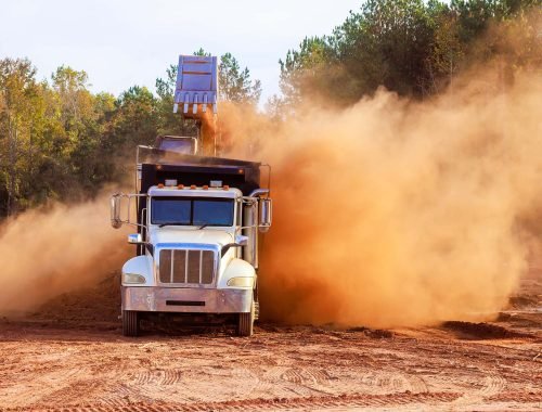 Construction truck is unloading materials at job site, sending cloud of dust into air is construction area surrounded by during land removal